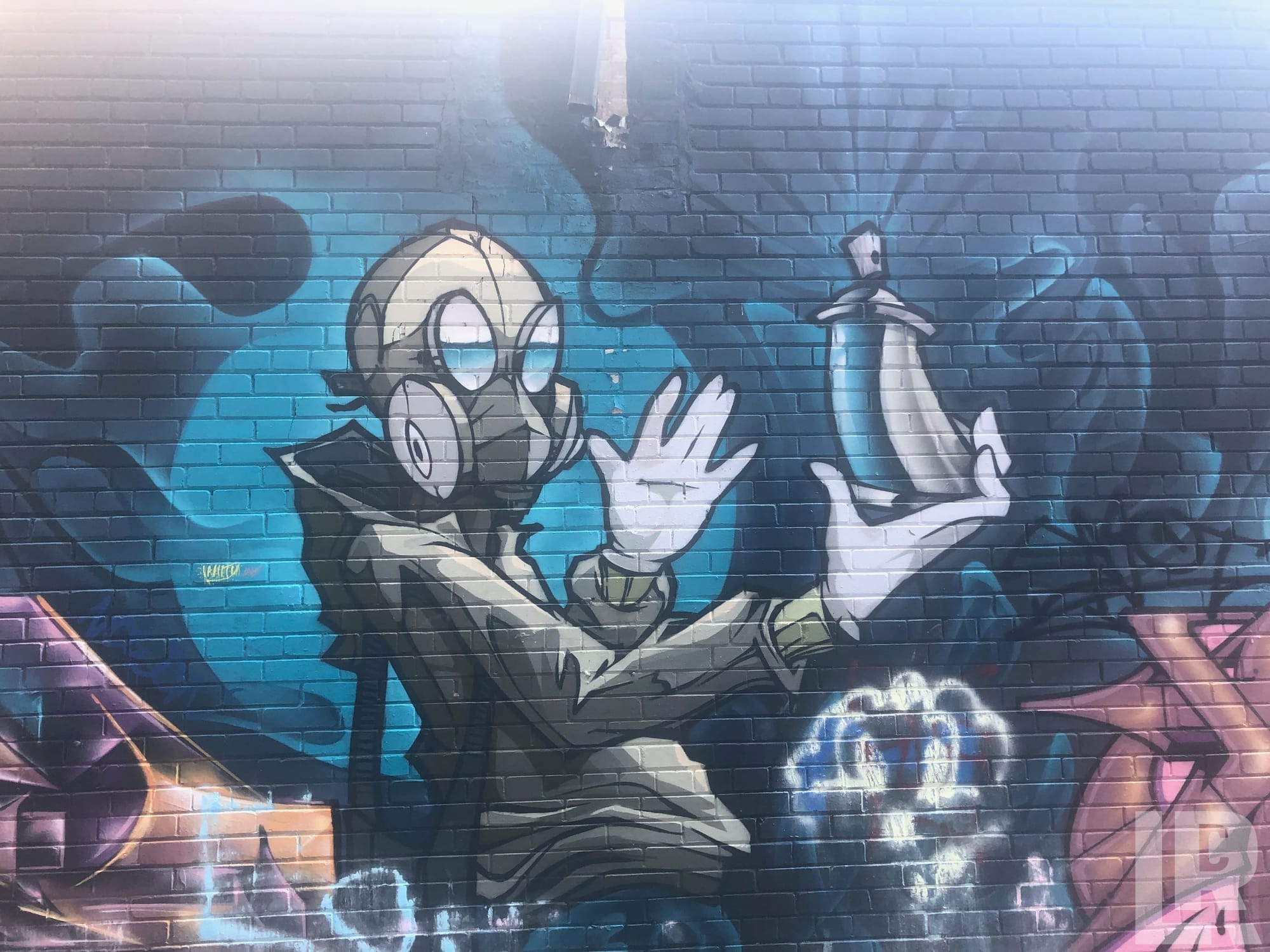 Street art mural of a gas-masked cartoon figure holding a spray can on a brick wall