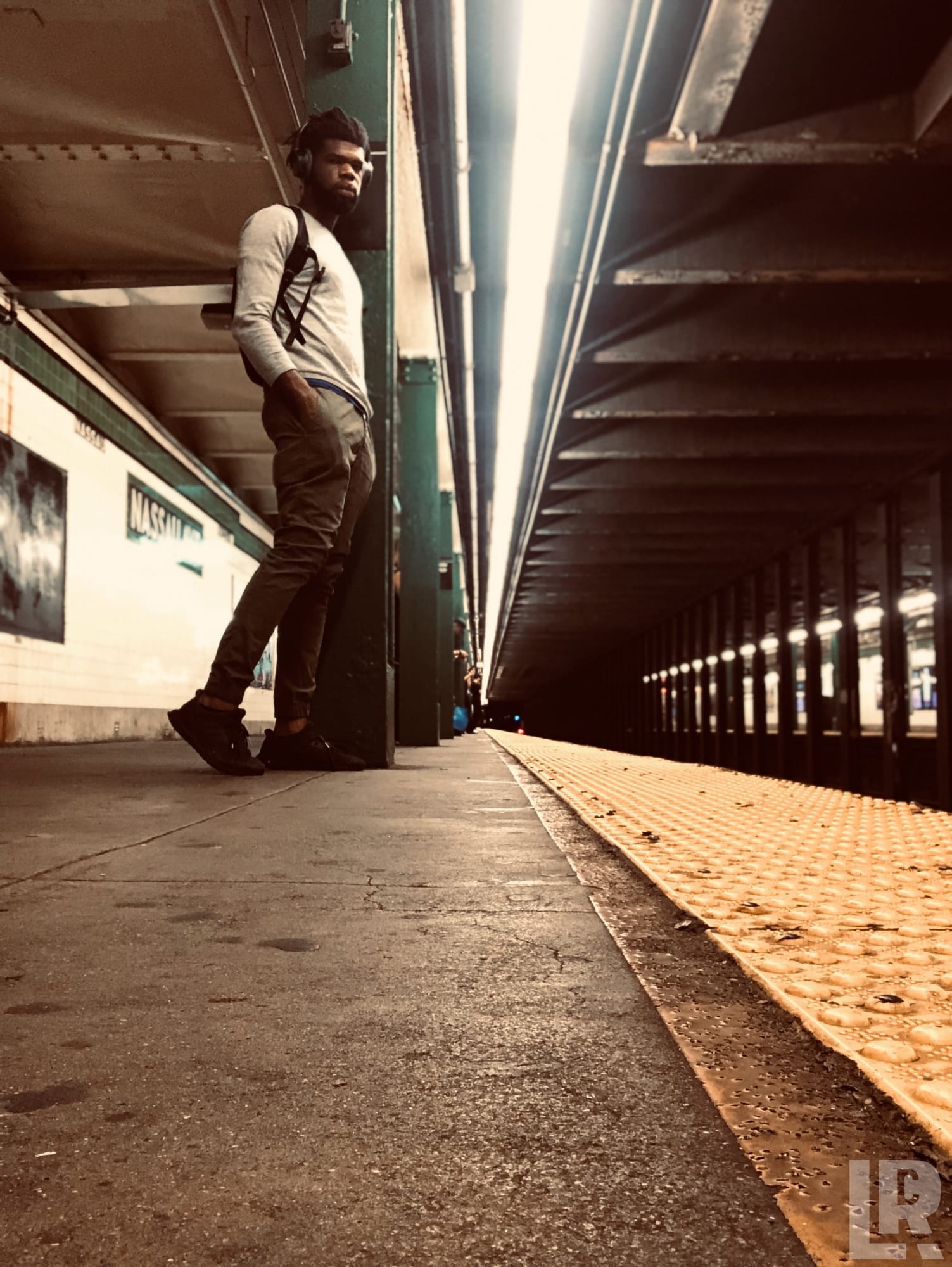 Man leaning against a subway platform pillar with headphones on
