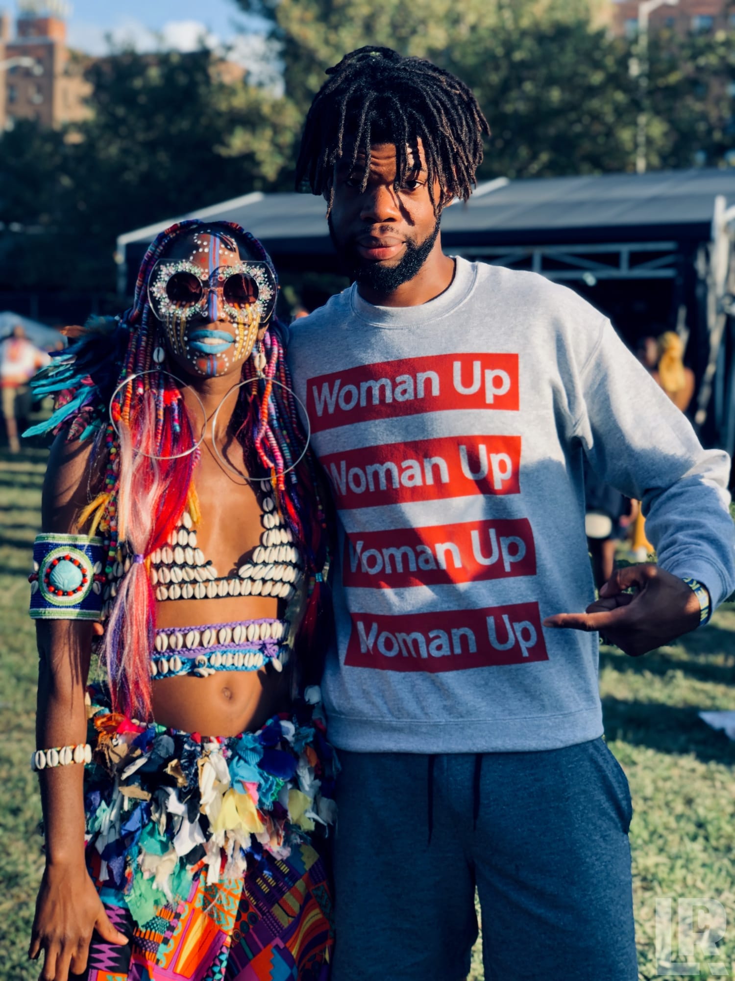 Two people at an outdoor festival, one in a colorful tribal costume with face paint, one in a Woman Up sweatshirt