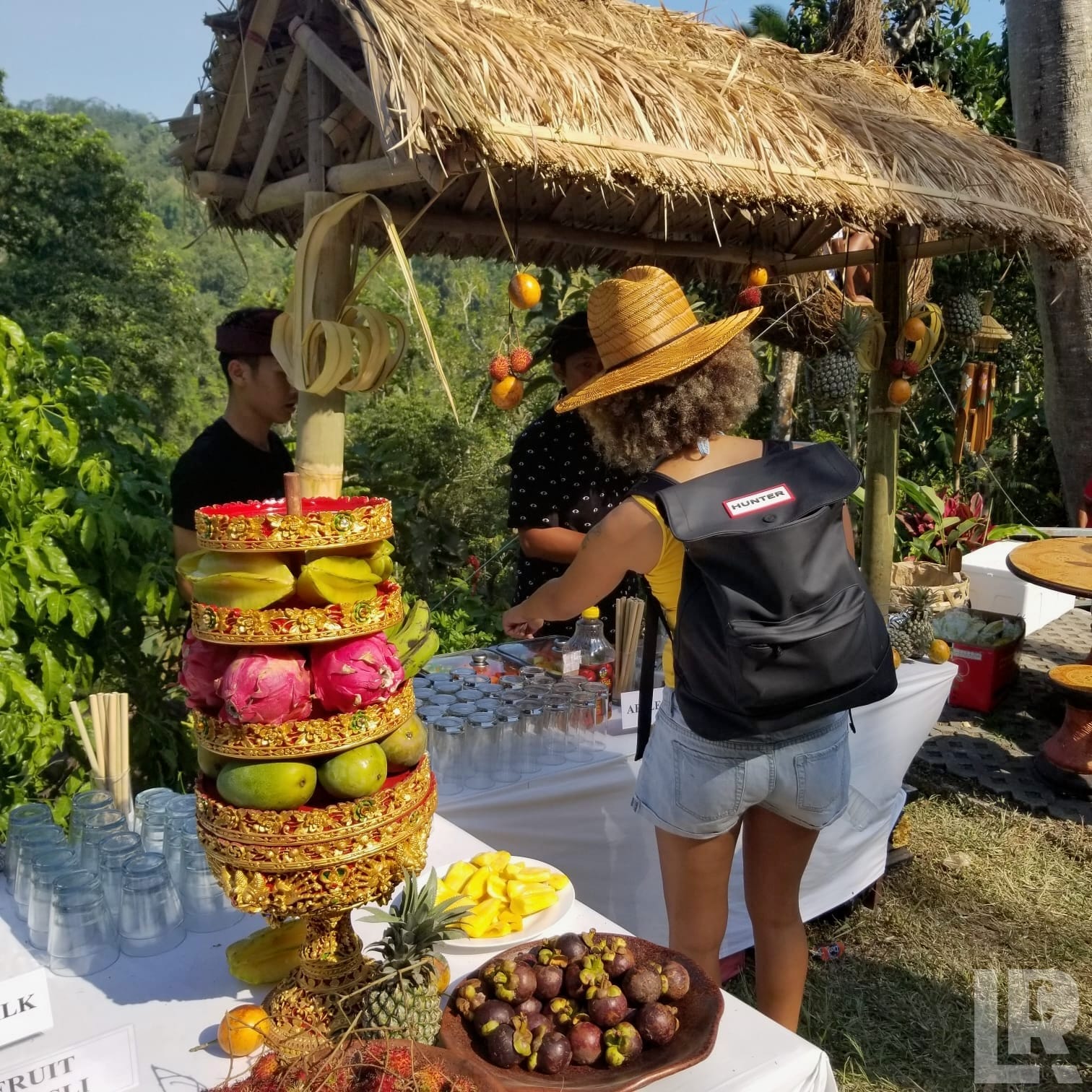 Woman at a tropical outdoor fruit buffet with tiered golden stand and thatched roof gazebo