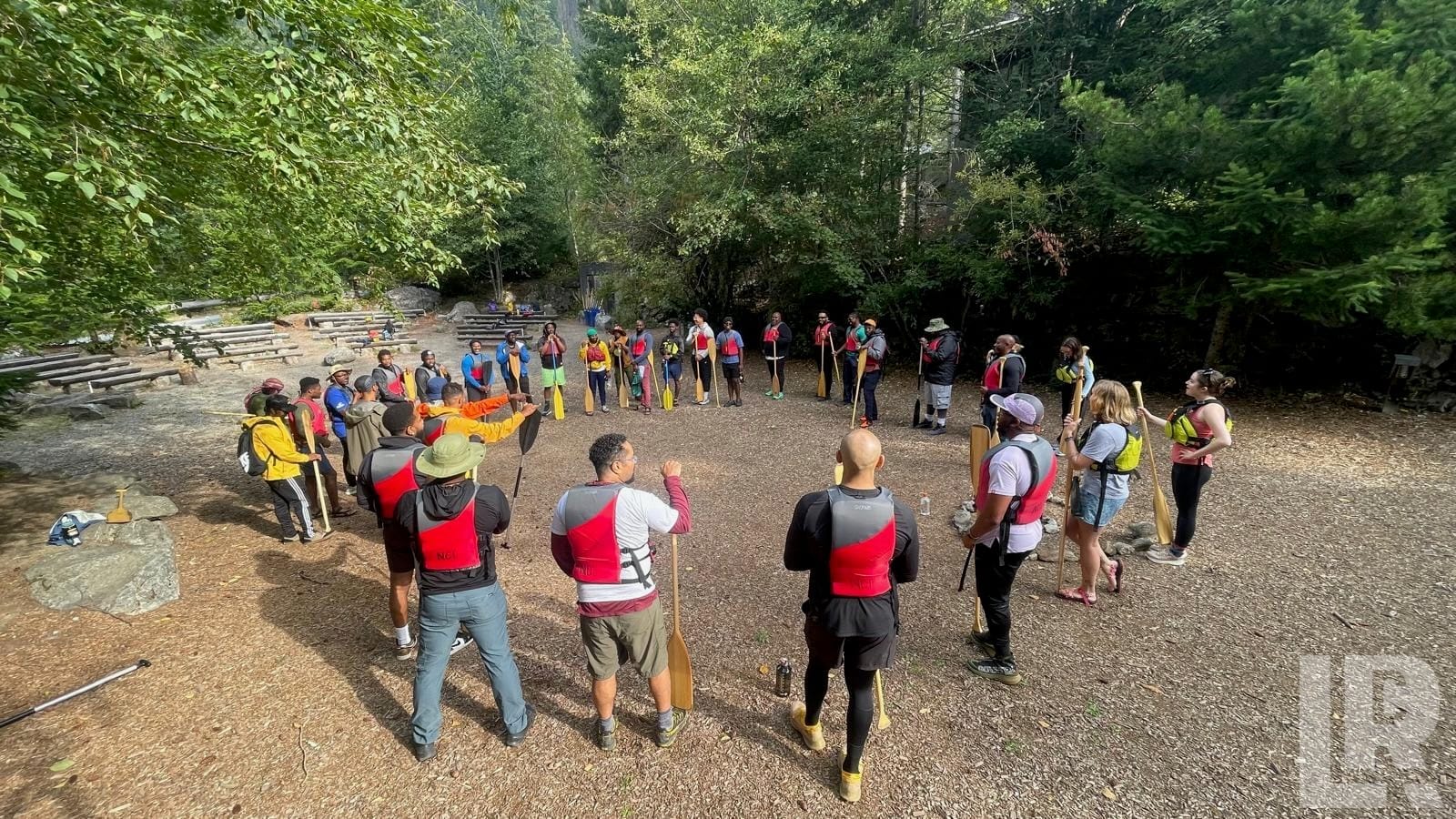 Group of adults in life vests holding canoe paddles gathered in a circle outdoors among trees